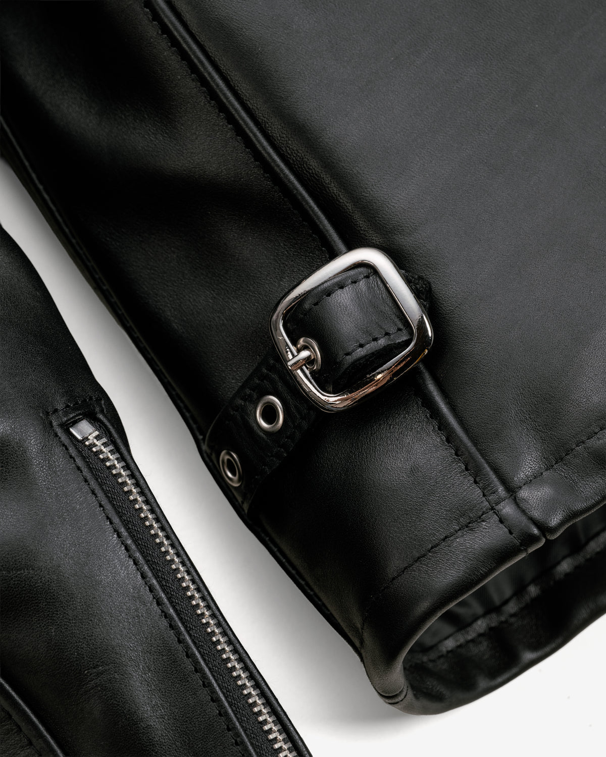 Close-up of a black leather jacket detail featuring a silver-toned metal buckle strap and a zipper, set against a plain white background.