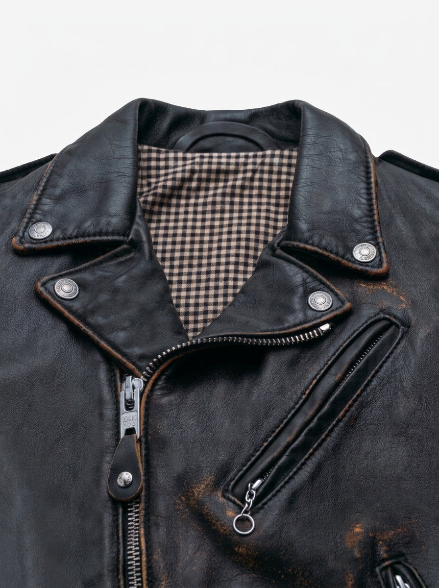 Close-up of a black distressed leather motorcycle jacket with silver snap buttons, a zippered chest pocket, and a brown and beige gingham patterned lining.