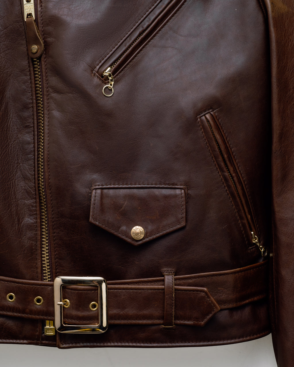 Close-up of a dark brown leather jacket with a matching leather belt, featuring brass hardware, a front zip closure, and a flap pocket with a snap closure.