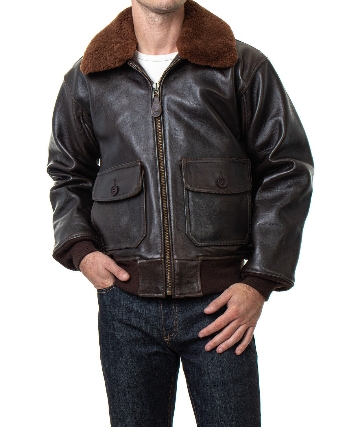 A man wearing a dark brown leather aviator jacket with a brown shearling collar, paired with a white t-shirt and dark blue jeans, shown from the neck down on a white background.