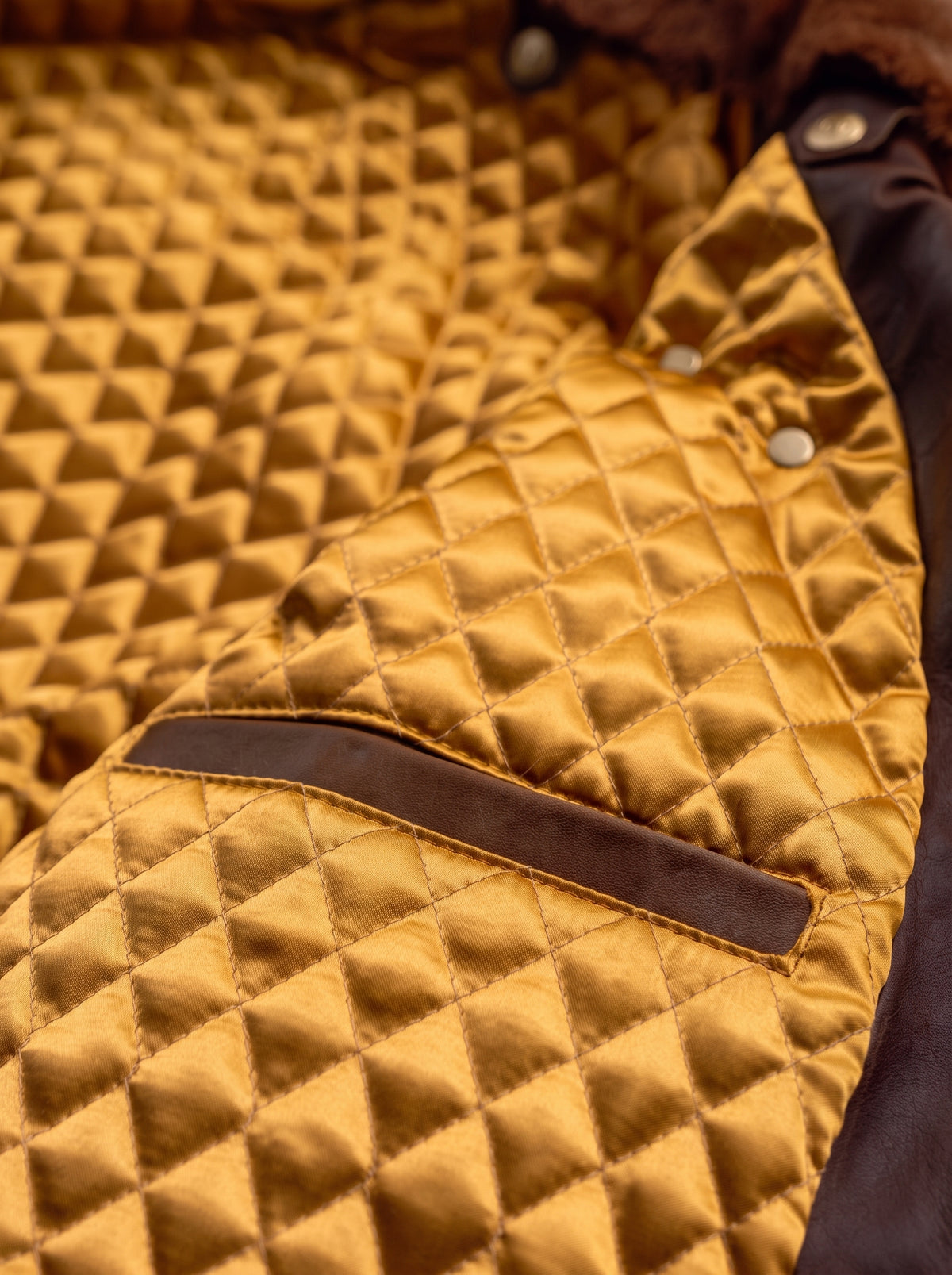 Close-up of a quilted mustard yellow jacket with a diamond-stitched pattern, a dark brown horizontal trim detail, and silver snap buttons.